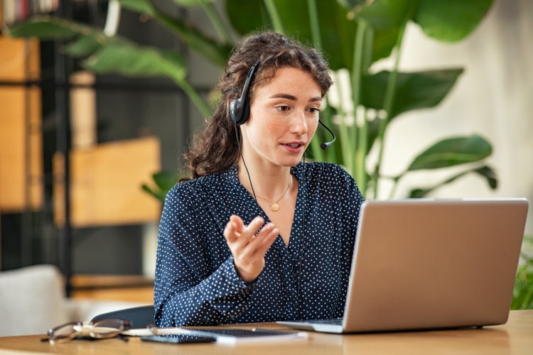 A woman wearing a headset with a microphone sits at a desk while managing a remote team from her laptop.