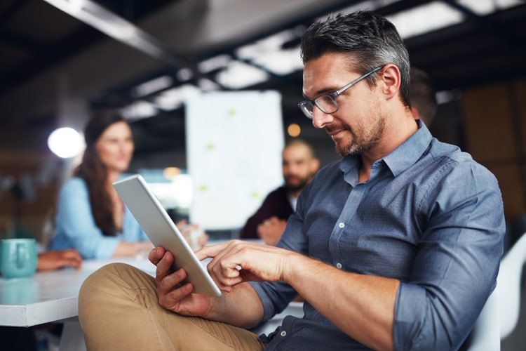 A man sitting on a chair holds a tablet to review an electronic document after his business transitioned to a paperless office