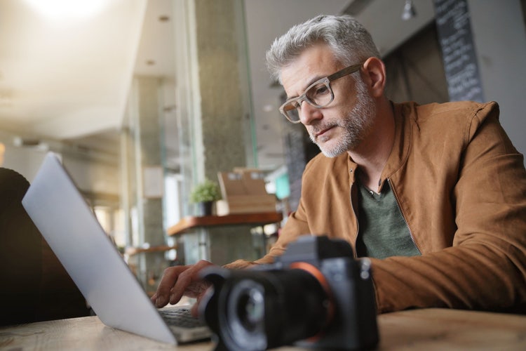 A man sitting at a table with a camera on it creates an invoice for photography on his laptop.