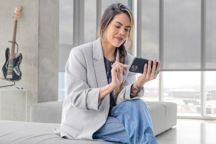 Woman in a blazer and jeans sitting on a gray bed in a luxury high-rise with a black electric guitar in the background organizing a digital cash receipt on a cell phone.