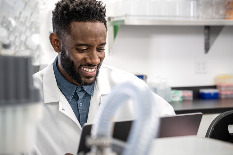 Person working in a lab wearing a lab coat and smiling while working on writing memo examples on his tablet to communicate with his entire staff.