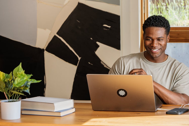 Man sitting at a wooden table with two books and a plant in a white pot, reading about RFI vs. RFP documents on a laptop.