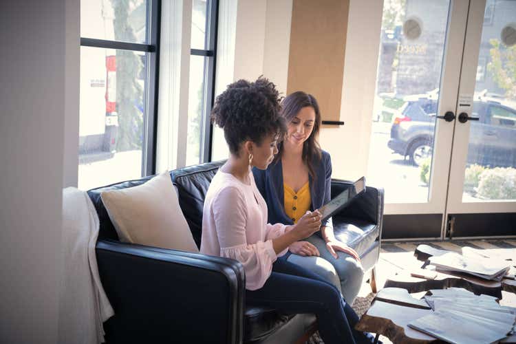 A woman sitting on a couch next to another woman electronically signs loan documents on a tablet.