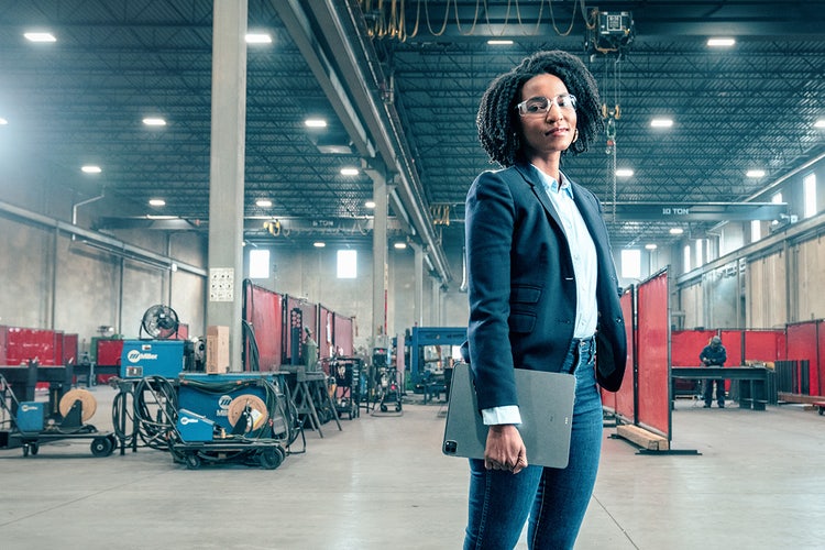 Woman in a suit jacket wearing safety glasses and holding a tablet that contains the weekly status report template is standing in front of an industrial workshop with welding equipment in the background.