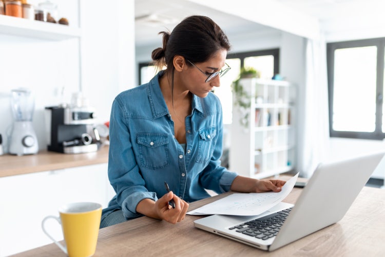 A woman sitting at a table looks at a piece of paper with a laptop in front of her.