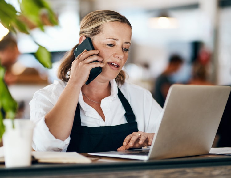 A woman sitting at a table and talking on her phone creates a catering invoice on her laptop.