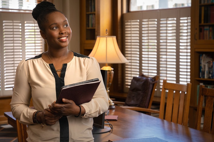 A woman stands in a meeting room with warm lighting and a large table with chairs, holding a laptop and notebook as she waits for people to arrive for a meeting that will leverage the board meeting agenda template she just created.