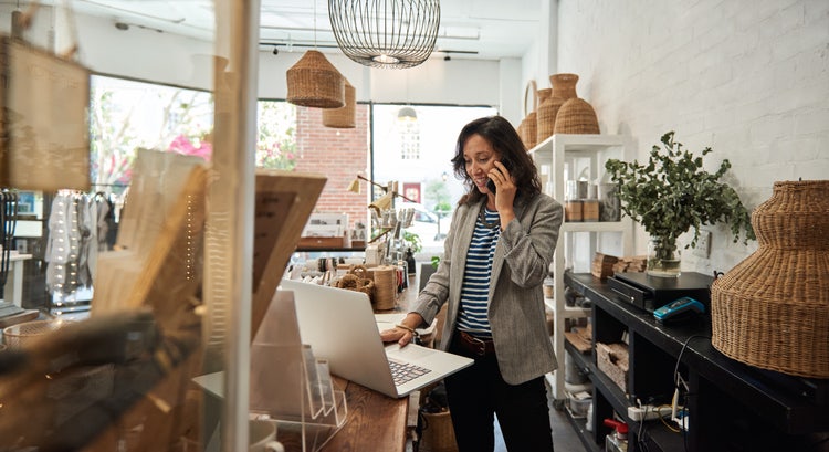 A woman standing in a retail business politely asks for invoice payment on her smartphone with her laptop in front of her.