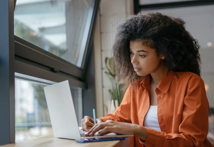 A woman sitting at a table writes on a piece of paper while reviewing her school's online program management software.