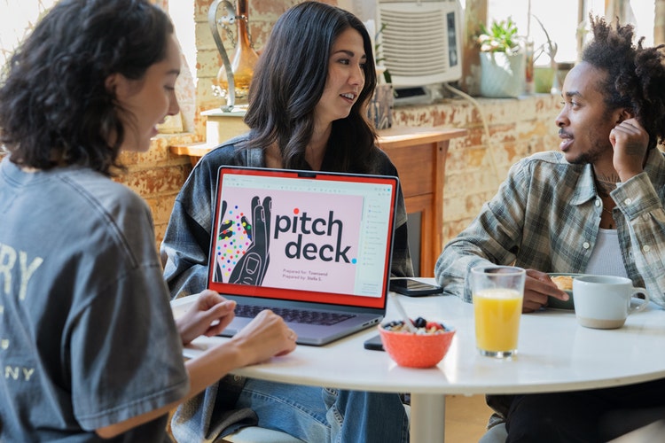 Three people sitting at a round, white table in a cafe having a light breakfast of oatmeal, orange juice, and coffee while working on a laptop learning how to create a sales pitch template.