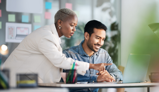 Woman and man collaborate on a project. Woman is standing, man is seated with a laptop.