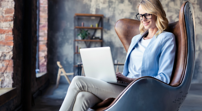 A woman with blonde curly hair and black glasses sits on a brown leather chair and reviews a loan agreement on her laptop computer.