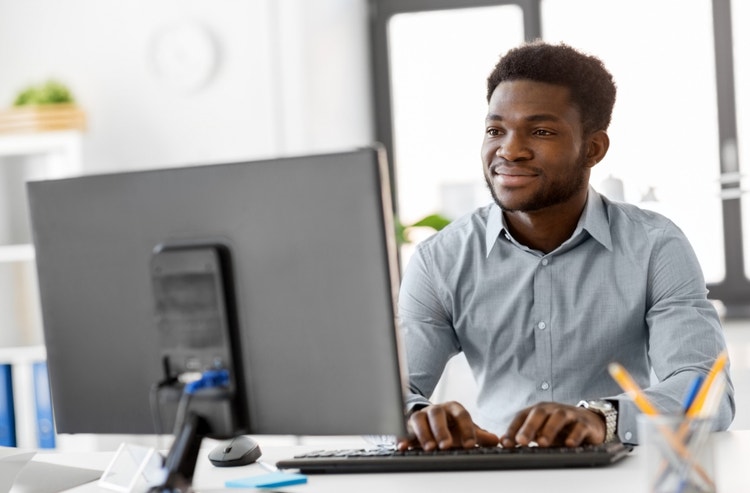 A man with dark skin and brown hair wearing a light blue button-down shirt sits behind a monitor and makes an invoice template in Google Docs.