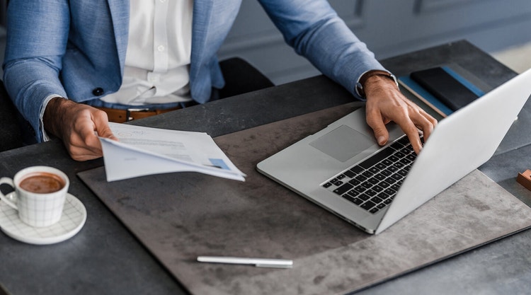 A man holding a contract in one hand and using his laptop computer at a desk with the other