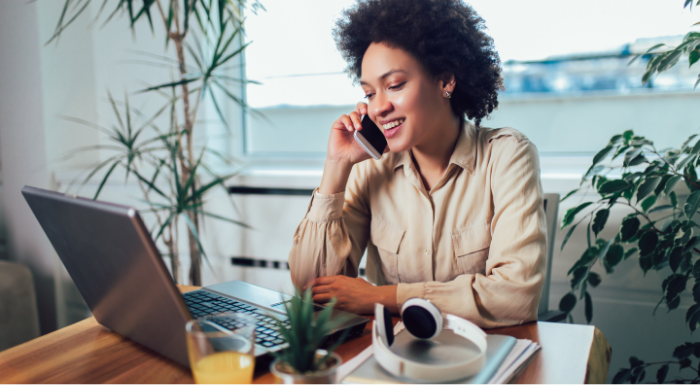 A woman sitting at a desk looks at her laptop and talks on her smartphone.