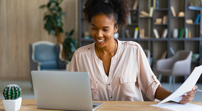 A woman sits at a desk with a laptop in front of her while holding a piece of paper.