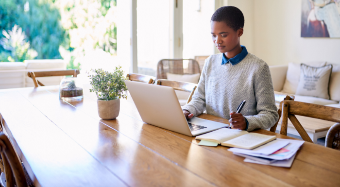 A man with dark skin and brown hair wearing a long-sleeved gray sweater with a blue collar sits at a long oak desk with a laptop and works on writing a loan agreement contract.