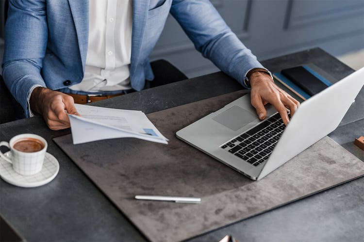 A man holding a contract in one hand and using his laptop computer at a desk with the other