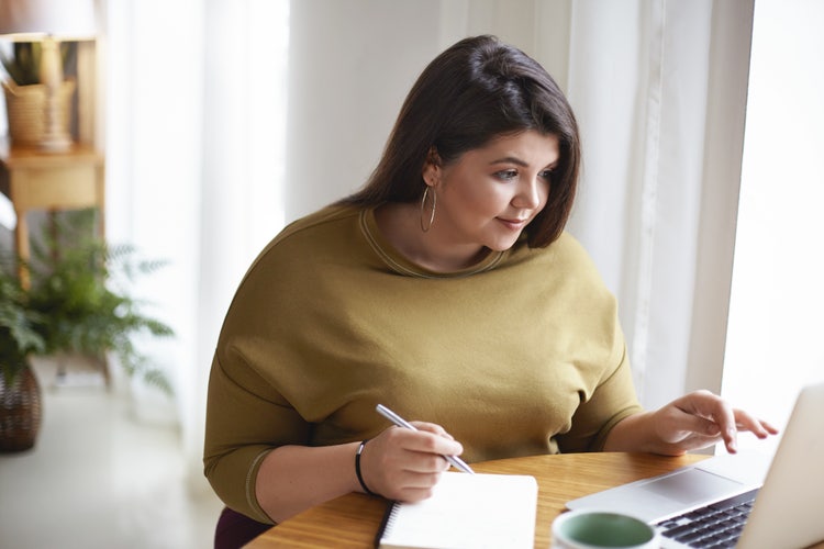 A woman sitting at a table looks at her laptop with a notepad sitting beside it and a pen in hand