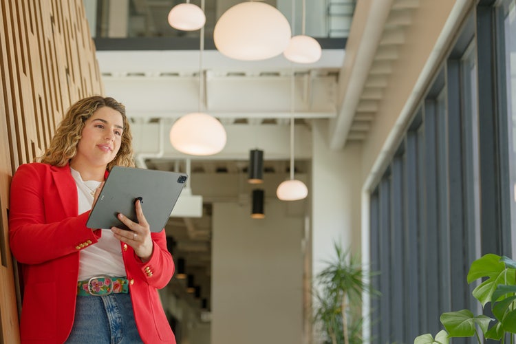 Person in an office hallway wearing a red blazer and bright belt working on a silver tablet, learning how to make a daily schedule.