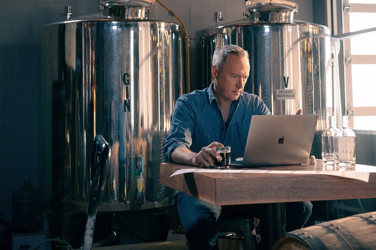 Person sitting at a wooden desk learning how to copy text from a secured PDF with metal distilling tanks labeled “Gin” in the background and wooden kegs in the foreground.