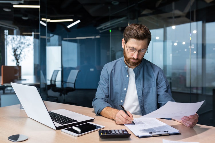 A man sitting at a table in an office writes out a cash flow budget on a piece of paper clipped to a clipboard.