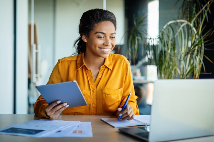 A project manager makes a tasks list at her desk with a pen, notebook, and laptop.