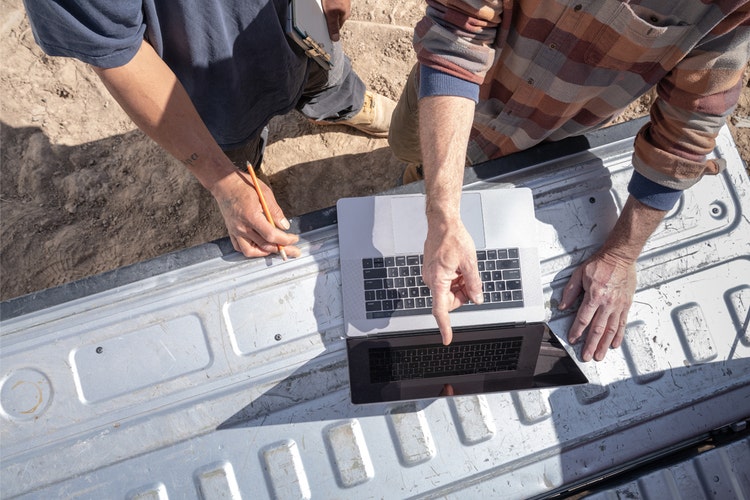 Two people working on a construction budget template using a computer sitting on the tailgate of a silver pickup truck.