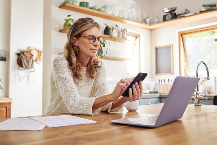 A woman sitting at a kitchen table uses her phone and laptop to digitize documents.