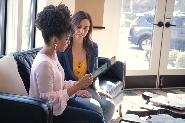 Two women sitting on a black couch with papers and books set out on the coffee table in front of them are reading about how to write an outline template for an essay on a tablet.