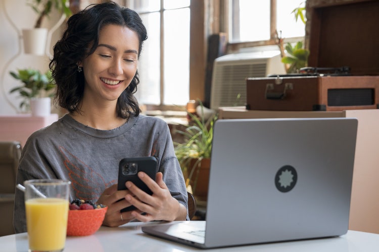 Person sitting in a kitchen, looking at their phone to read a friend reference letter, with a glass of orange juice, a bowl of fruit, and a laptop sitting on the table, a wooden record player on a table behind them, and surrounded by green plants.