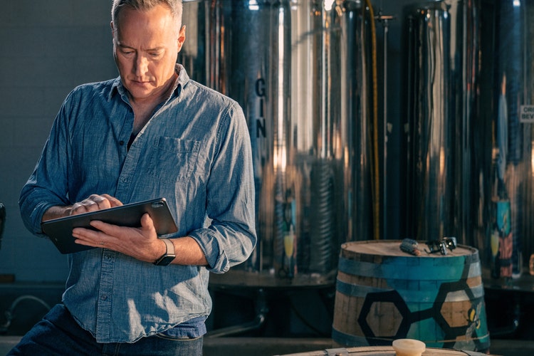 A person in a blue shirt standing in front of distilling tanks and barrels using a tablet to make a job description template to hire for the business.