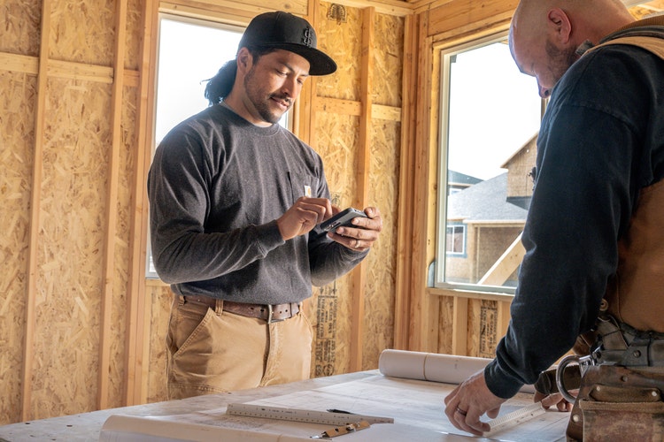 Two people at a construction site in an unfinished house looking over plans with one person creating a note taking template PDF on a cell phone.