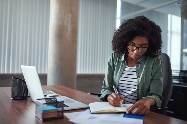 A woman in an office uses a laptop and a notebook to organize work tasks with a work to-do list.