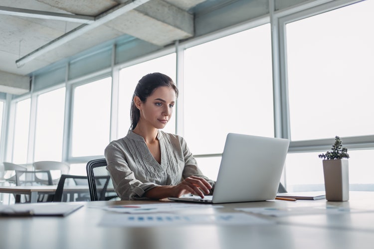 A woman sitting at a table redlines a document before signing it on her laptop.