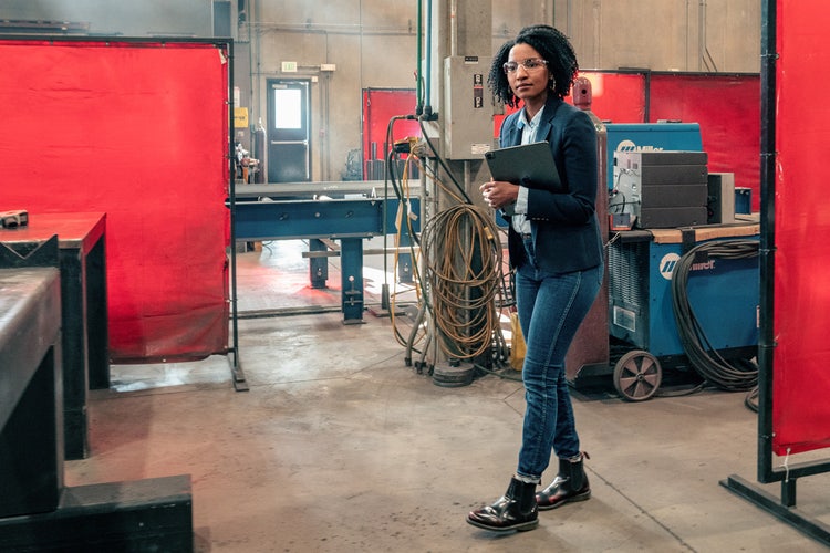 Person wearing a blue blazer, jeans, and safety glasses, walking through a welding shop holding a tablet that has a summary of a long-form document created by a PDF AI summarizer.