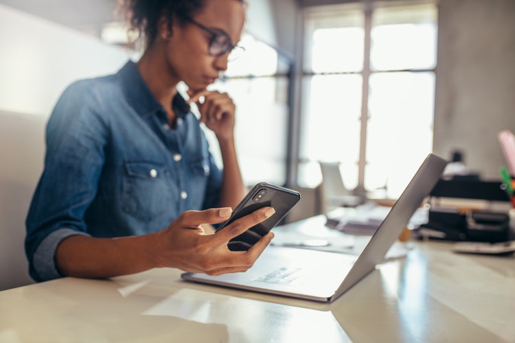 A woman holding a phone looks at her laptop while creating a mobile-friendly PDF.