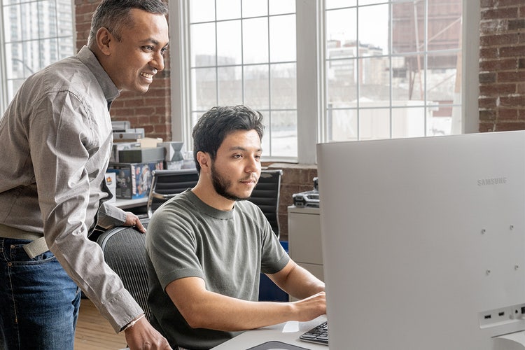 Two men looking at a computer screen in an office with large windows and brick walls as they use an AI Assistant to ask PDF questions.