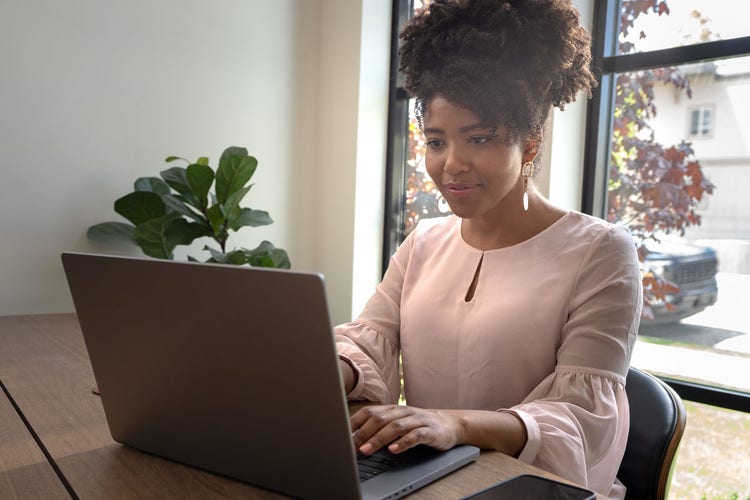 Person in a pink shirt and long earrings sitting in front of a window next to a green plant working on a laptop to learn how to fix a PDF.