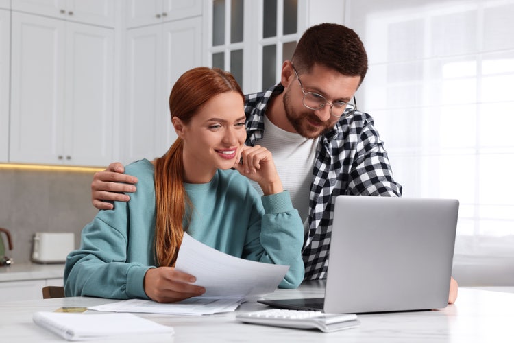 A man and a woman sitting at a kitchen table explore what they need for a loan approval on their laptop.