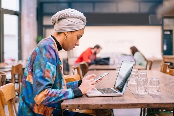 A person sitting at a table using their laptop while looking at their cell phone