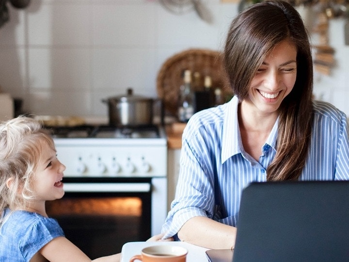 A photo of a parent working on their laptop at a kitchen table as their child tries to get their attention