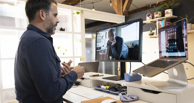 A person standing at their desk having a virtual meeting with another person using their laptop connected to a desktop monitor