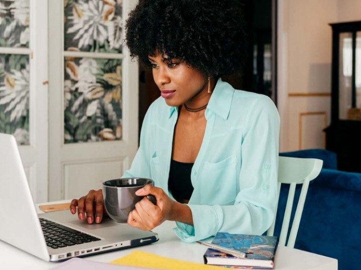 A woman in a blue shirt holding a cup of coffee working on a to-do list on her laptop.