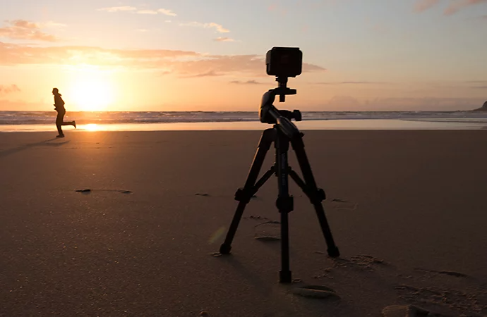 A camera on a tripod pointing at a person running on a beach with the sun setting in the background