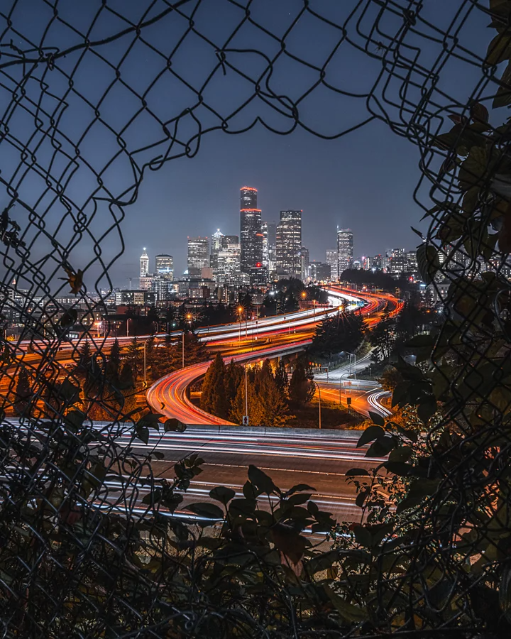 Light trails of a highway with a lit-up cityscape in the background seen through an opening of a gated fence