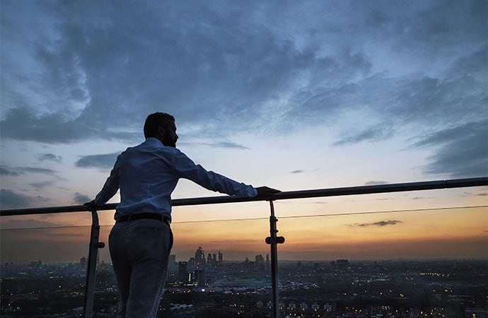 Man standing on balcony facing a city skyline at sunset.