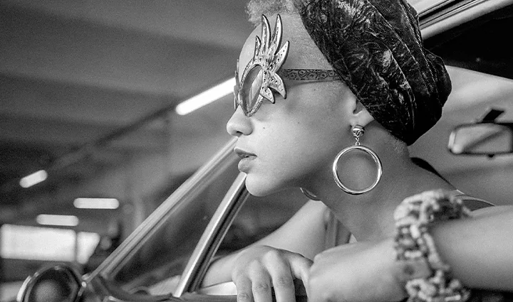 A monochrome photo of a person wearing vintage sunglasses sticking their head out of a car window