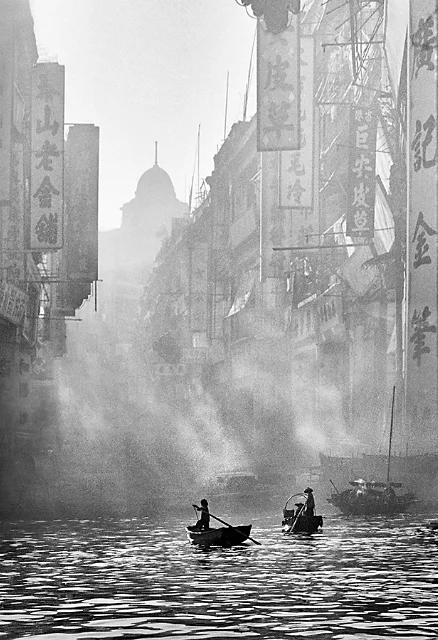 Monochrome photo of two small boats in water navigating between buildings