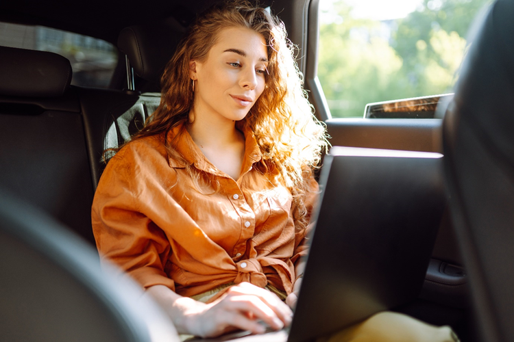 Photo of a woman sitting in the back seat of a car working on a laptop.